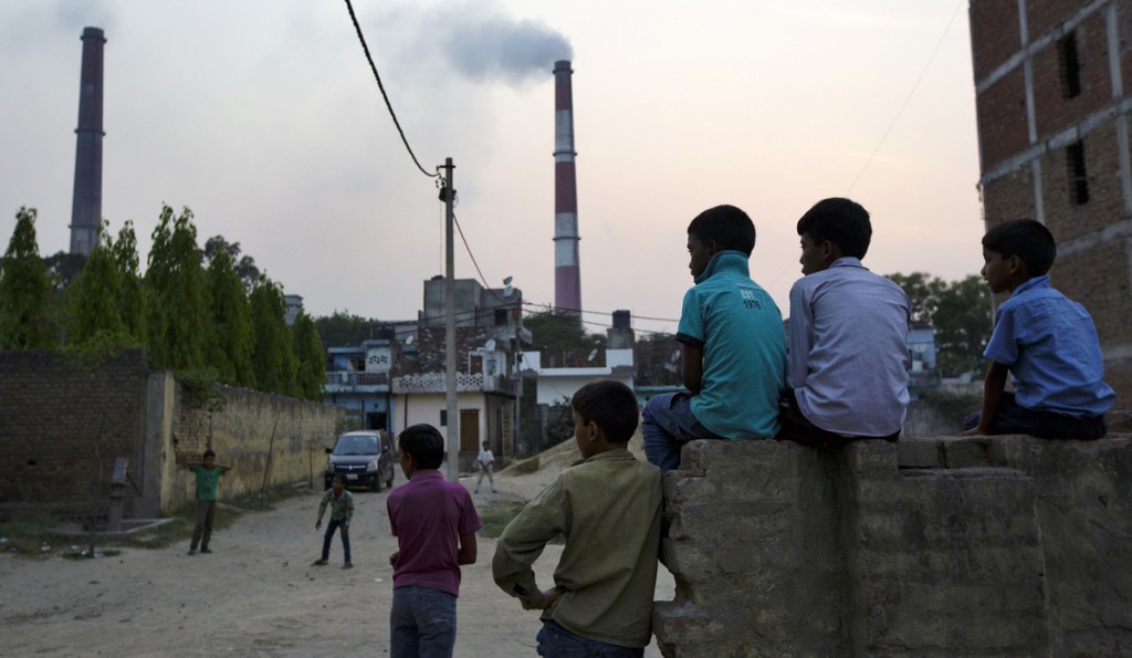 Boys sit watching street cricket as emissions billow from smokestacks in Badarpur, India. Photographer: Bloomberg