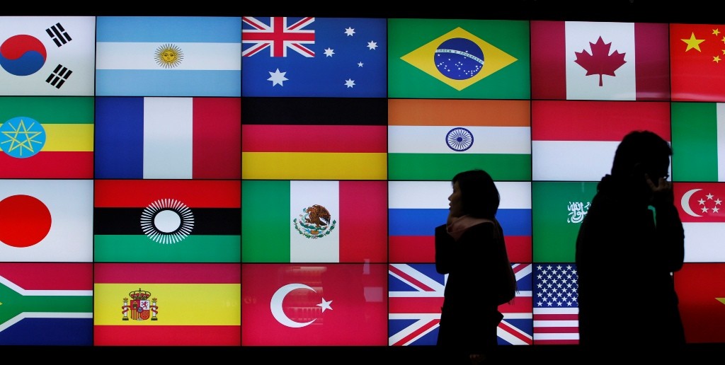 People using their mobile phones walk past a screen showing flags of the participating countries in the upcoming G20 summit in Seoul November 5, 2010. China on Friday pushed back strongly against U.S. policies ahead of a G20 summit, ridiculing Washington's plan to set current account targets and warning of risks in the Fed's monetary easing. REUTERS/Jo Yong-Hak